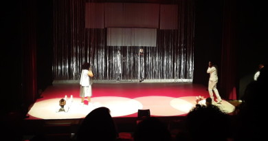 Em um palco de teatro, um homem vestido de branco está deitado, com as mãos no rosto. Uma mulher, de costas para ele, também com as mãos no rosto olha para outro homem, também vestido de branco.