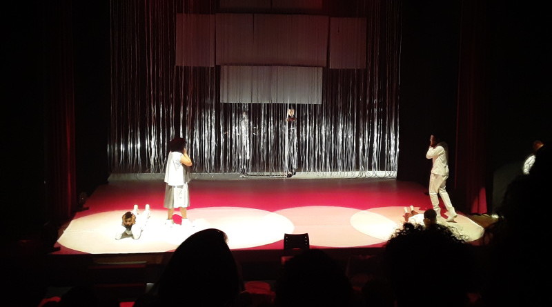 Em um palco de teatro, um homem vestido de branco está deitado, com as mãos no rosto. Uma mulher, de costas para ele, também com as mãos no rosto olha para outro homem, também vestido de branco.