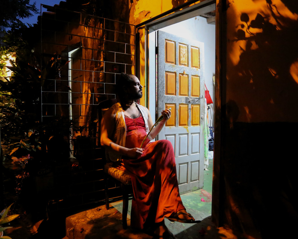 #pracegover: Foto noturna, sombras de plantas e casas ao fundo. No centro da imagem sentada de perfil, Yuri Tripodi, mulher branca de barba e cabelos castanhos está encostada na grade da porta de sua casa, ela veste vestido vermelho e colete branco. Segura uma planta, espada de Oxossi, nas mãos e está de pernas cruzadas e sandália. Seu rosto é iluminado pela luz que vem de dentro da casa, que está de porta aberta. A porta é azul clara, com nove quadrados amarelos pintados. A casa por dentro tem a parede azul e o chão verde.